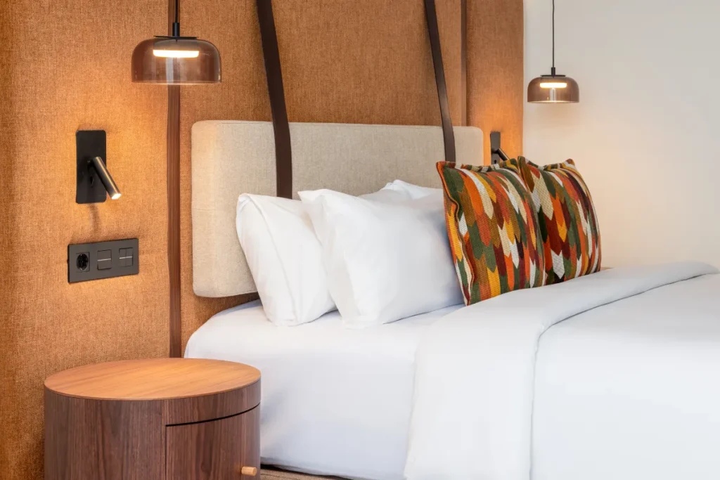 Cama con cojines blancos y almohada estampada, l&aacute;mparas y paredes de tela.