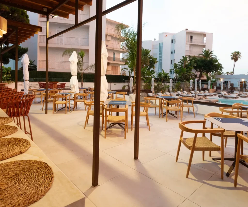 Terraza de hotel con sillas de madera, mesas y camas junto a la piscina.