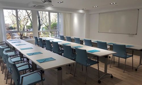Aula moderna con mesas, sillas azules y un pizarr&oacute;n blanco en la pared.