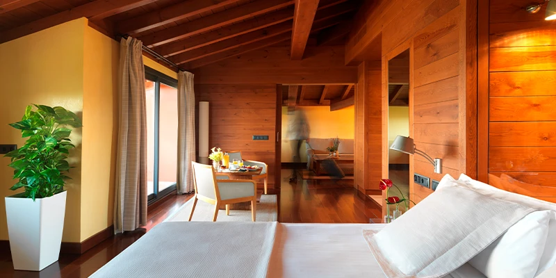 Habitaci&oacute;n r&uacute;stica con techo de madera, cama y mesa con sillas junto a ventana.