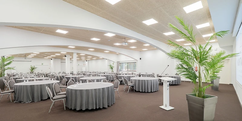 Sala de conferencias con mesas y sillas, plantas y podio al frente.