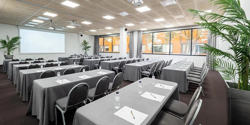 Sala de conferencias vac&iacute;a con mesas grises y sillas, decorada con plantas.
