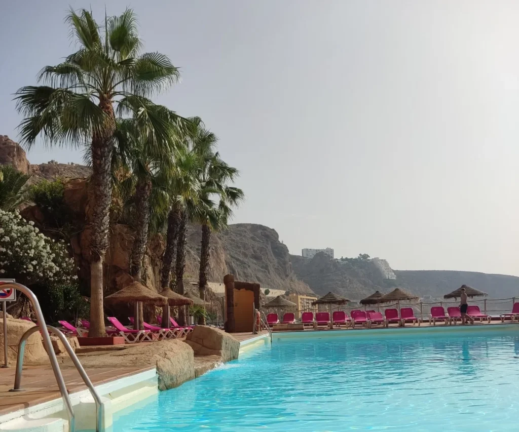 Piscina con tumbonas moradas y palmeras junto al mar y acantilados