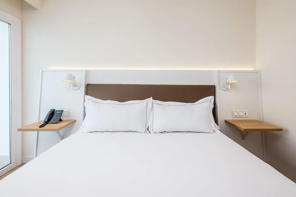 Habitaci&oacute;n moderna con cama doble, cojines blancos y mesas laterales.