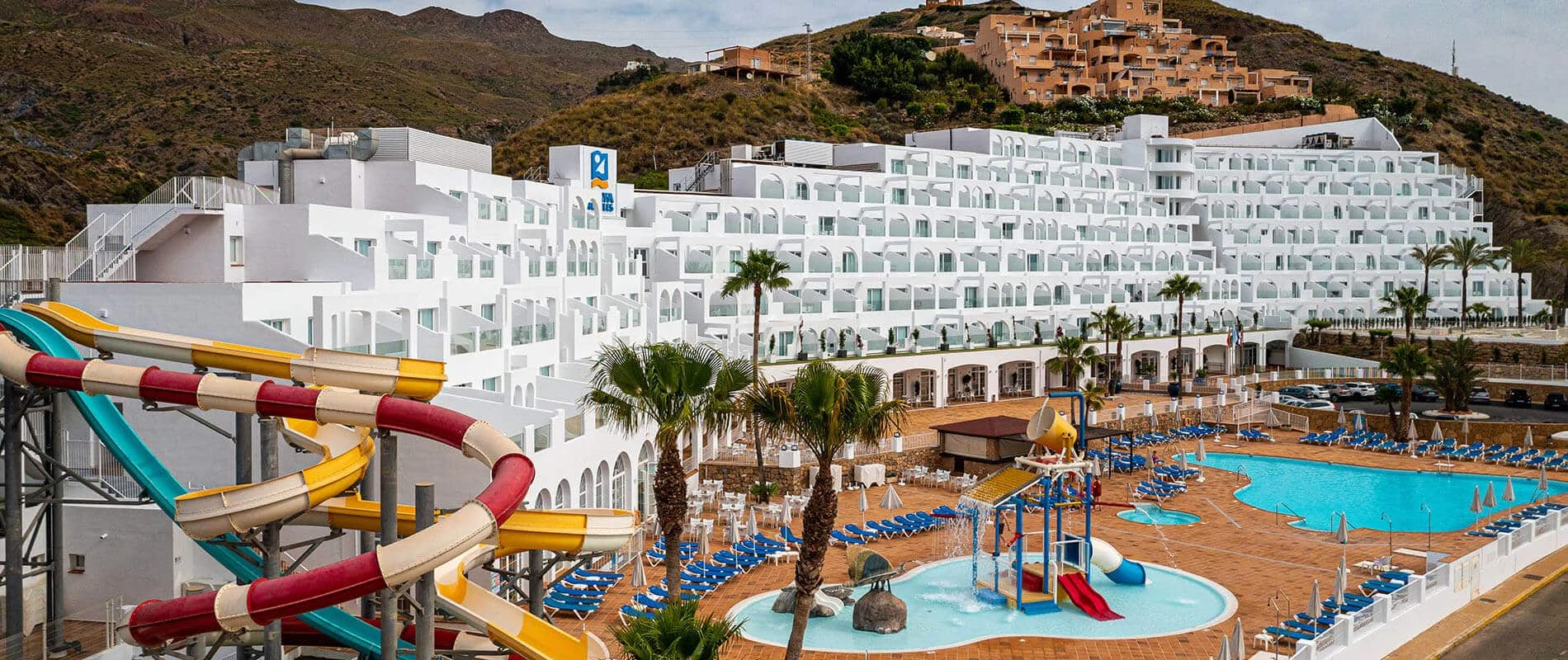 Hotel con piscina y toboganes de colores frente a monta&ntilde;as y ladera.