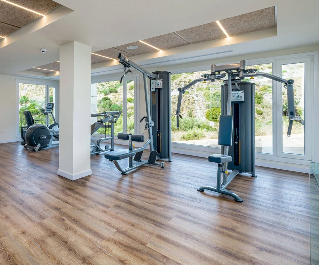 Gimnasio moderno con m&aacute;quinas de ejercicio y ventanales frente a jard&iacute;n.