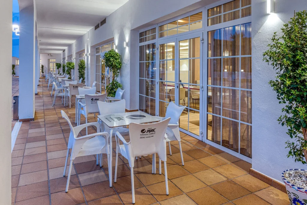 Terraza con mesas y sillas blancas junto a ventanales, con plantas decorativas