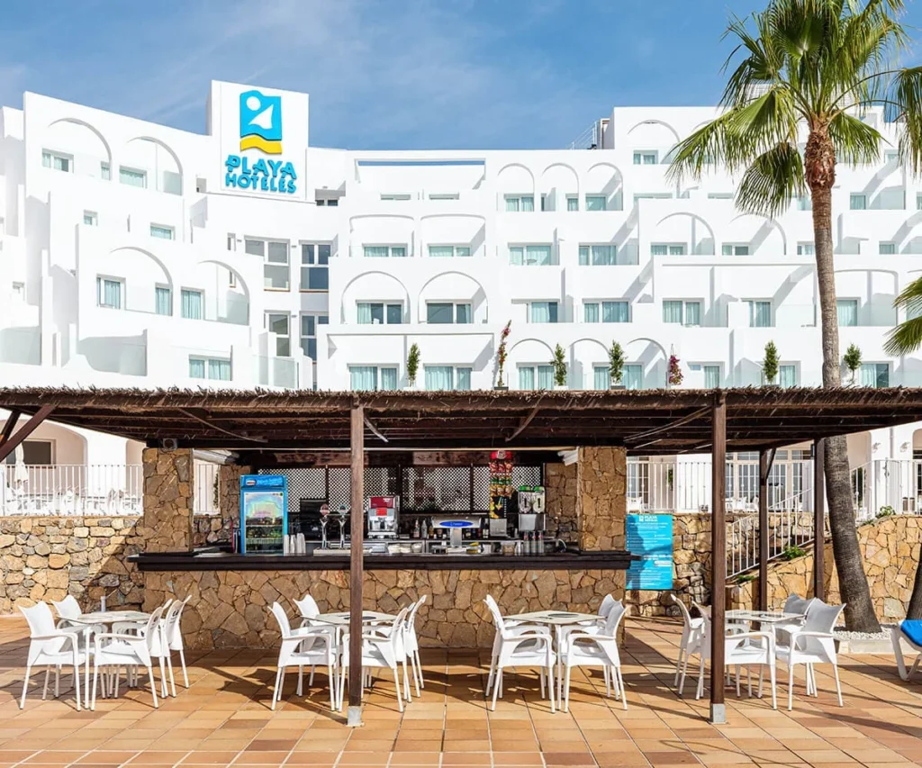Bar de hotel &ldquo;Playa Hoteles&rdquo; con palmera y sillas blancas al aire libre.