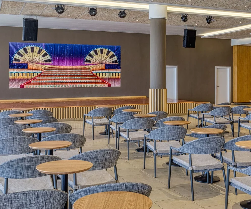 Sala de conferencias con sillas azules y mesa, escenario con pantalla art&iacute;stica