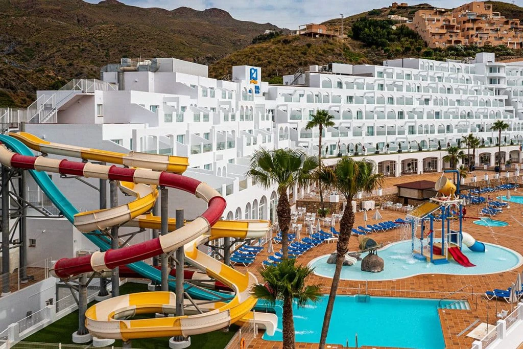 Piscina y toboganes multicolor en hotel con montañas al fondo.