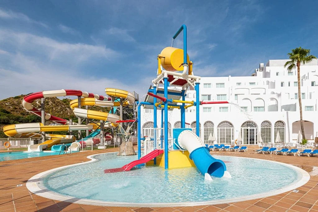 Parque acuático con toboganes y piscina circular frente a un hotel blanco