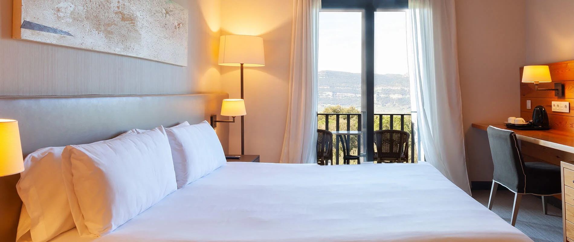 Habitaci&oacute;n de hotel con cama blanca, l&aacute;mparas y balc&oacute;n con vista al paisaje