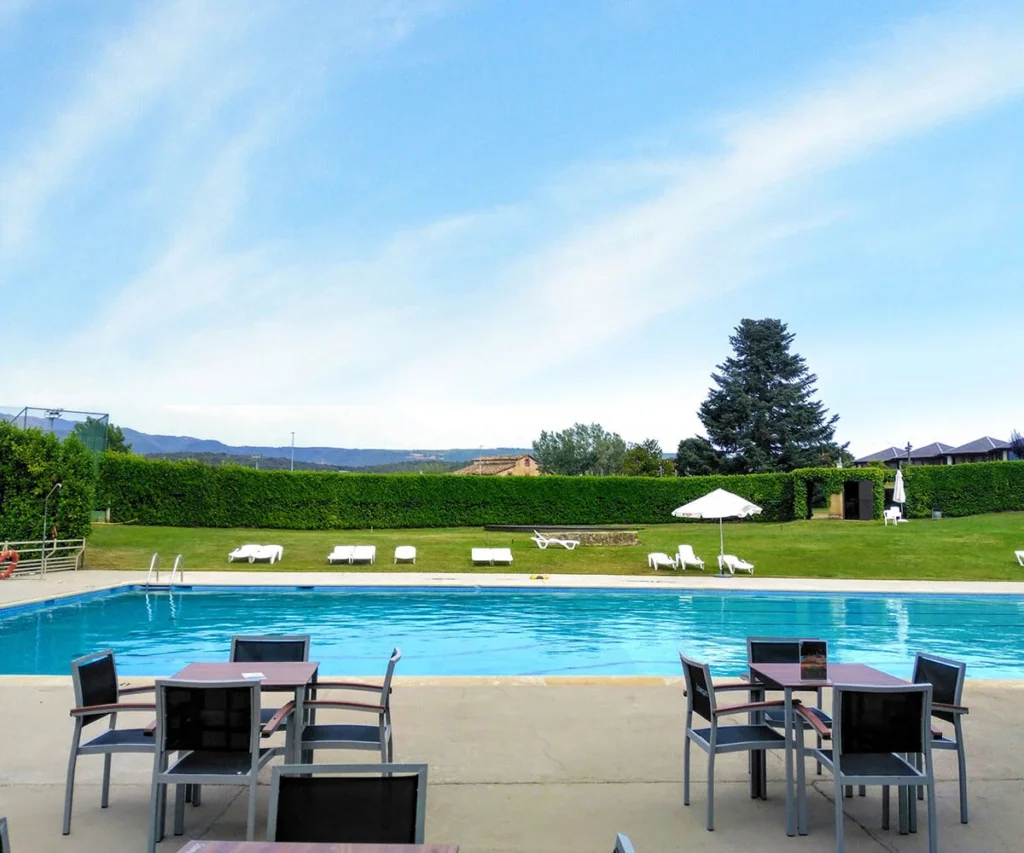 Piscina con sillas y sombrillas en un jard&iacute;n, cielo azul y nubes.