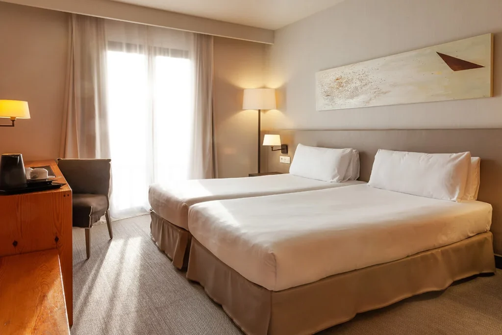 Habitaci&oacute;n de hotel con cama doble, mesita y l&aacute;mparas junto a ventana