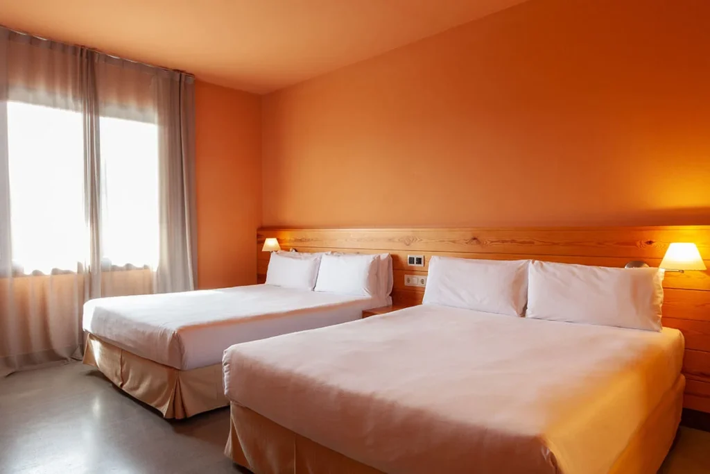 Habitaci&oacute;n de hotel con dos camas y luz c&aacute;lida, cortinas y pared naranja.