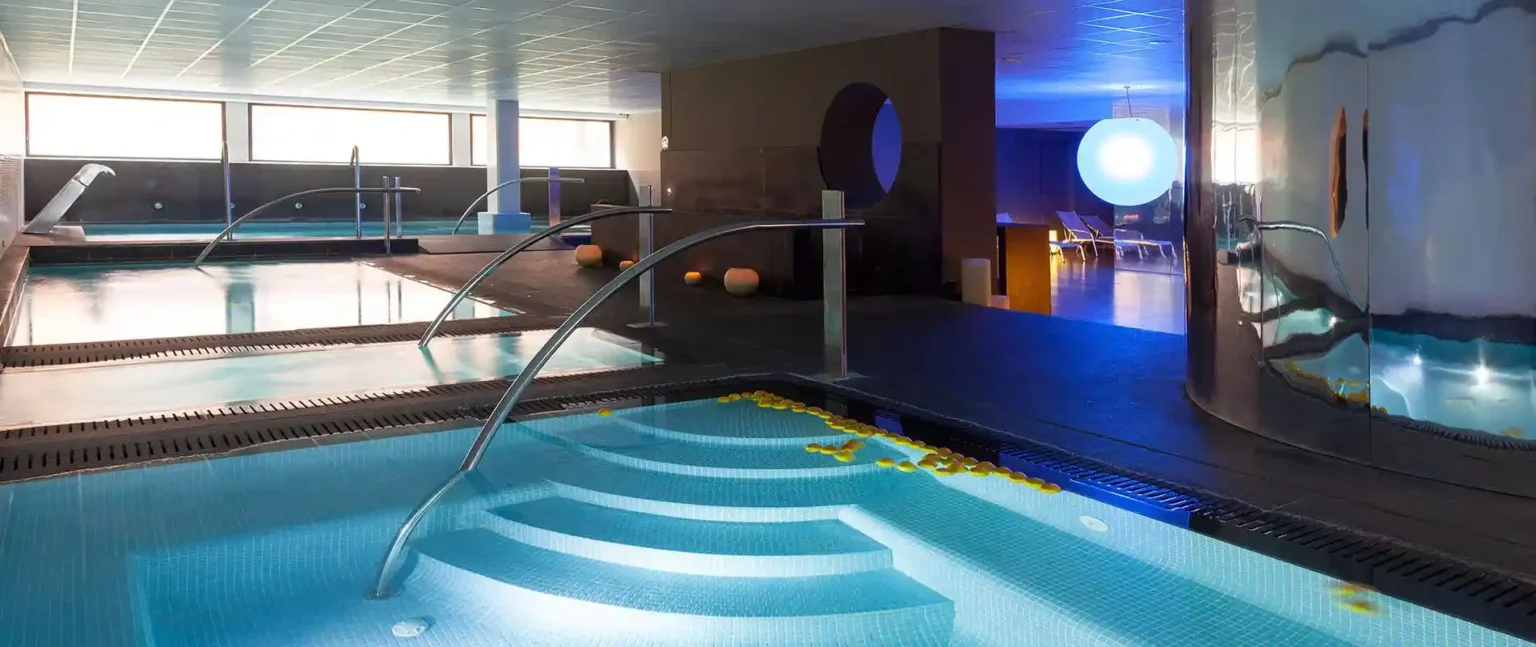 Piscina cubierta moderna con pasarela, luces y zona de hidromasaje.