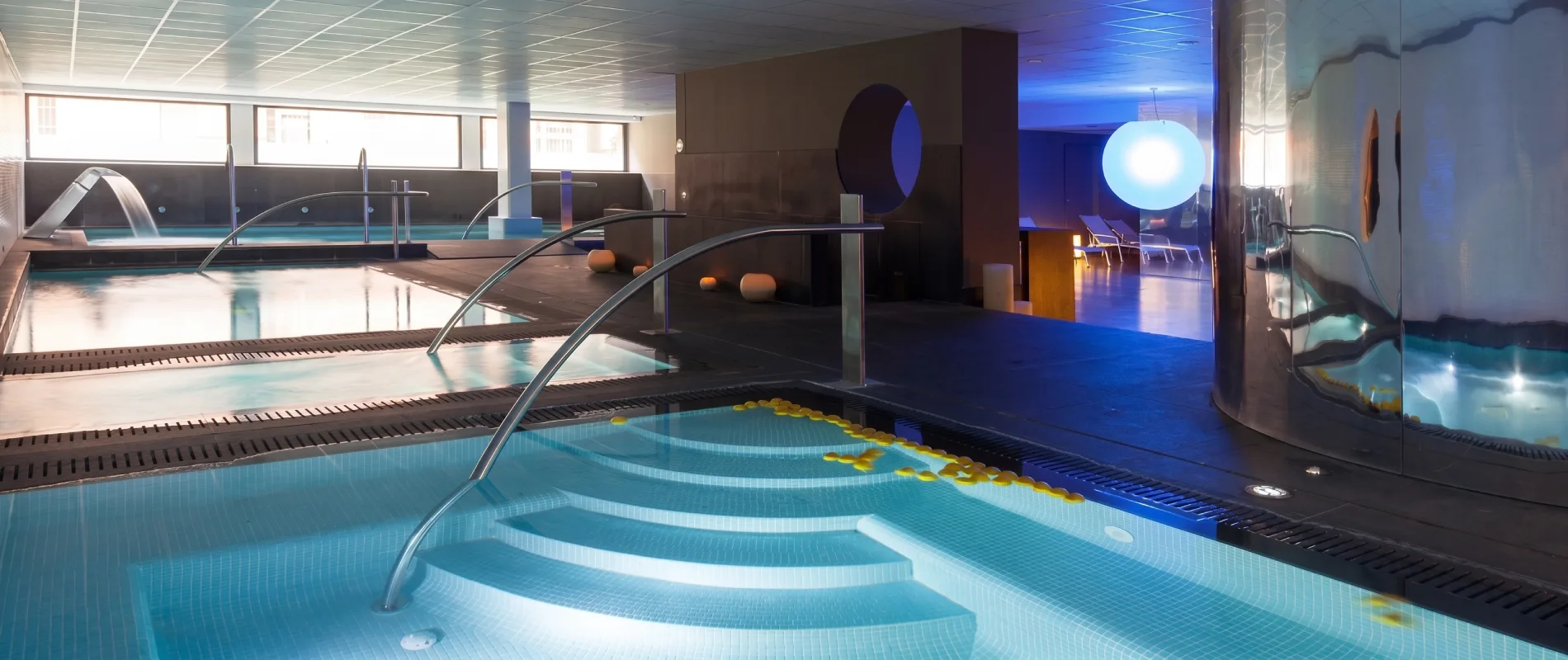 Piscina cubierta moderna con pasarela, luces y zona de hidromasaje.