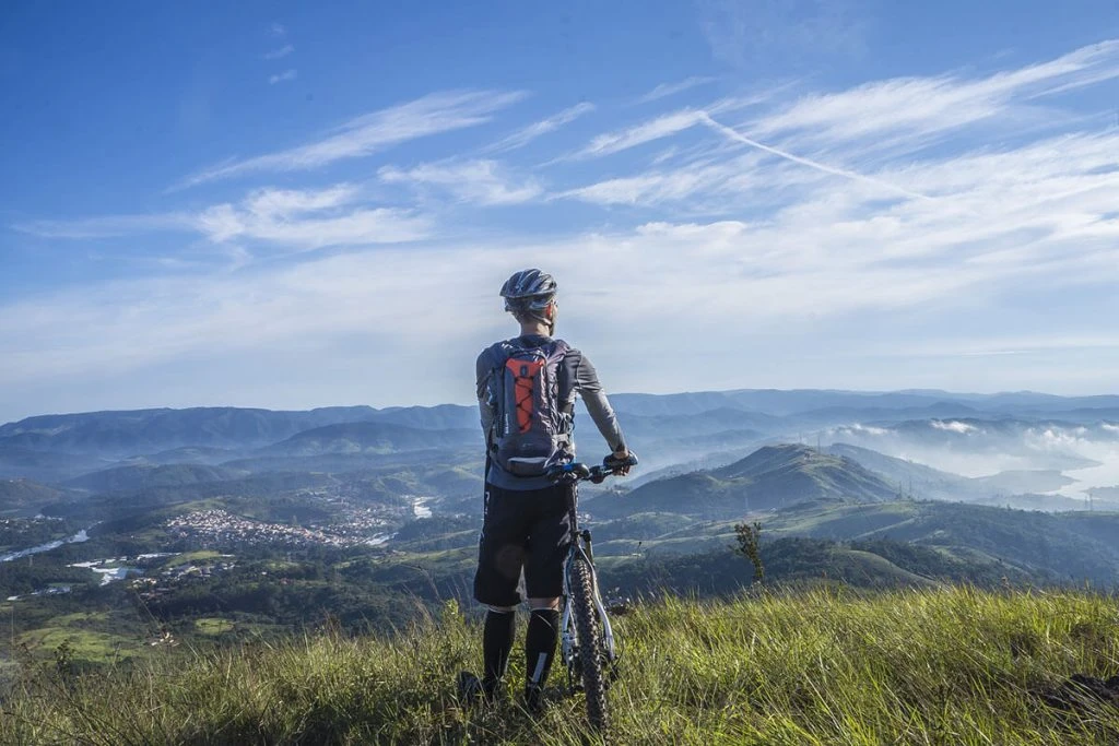 Ciclista con mochila y bicicleta en una colina, paisaje de monta&ntilde;as y lago al fondo.