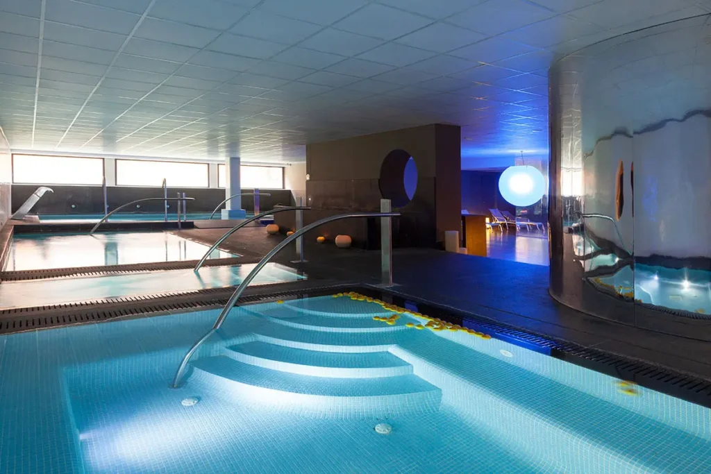 Piscina climatizada interior con chorros y luces azules, sala de spa.