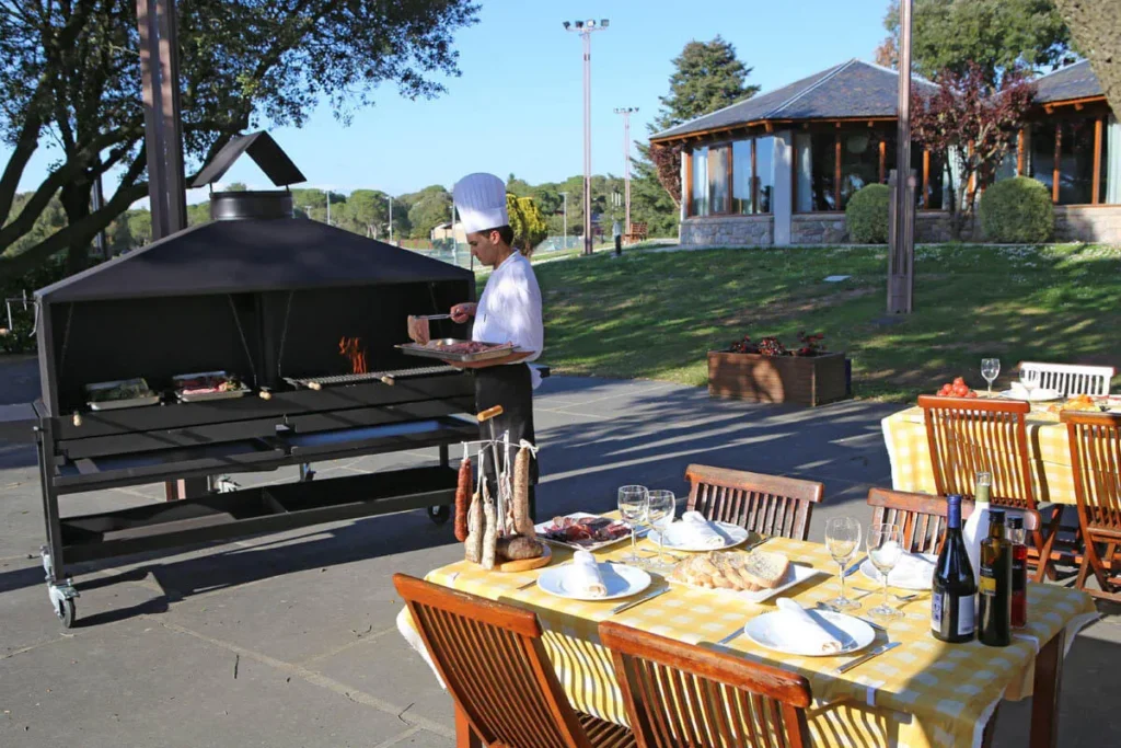 Chef con gorro cocinando en parrilla, mesa puesta al aire libre con vino y copas