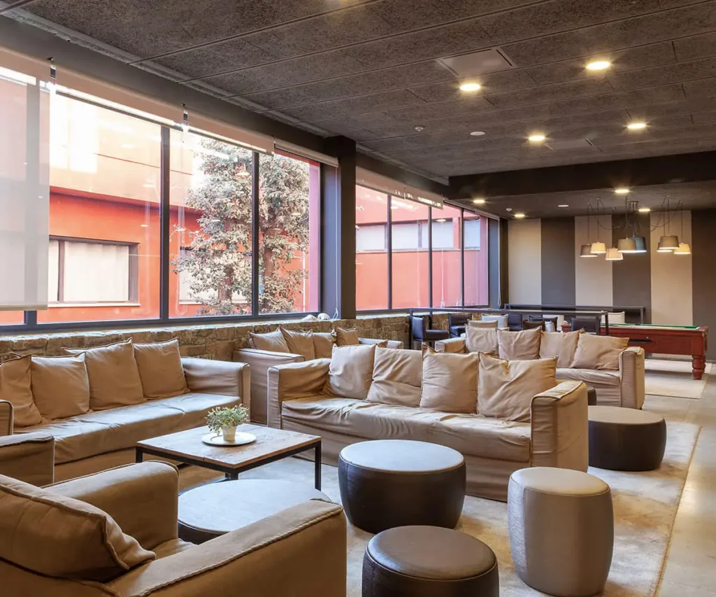 Sala moderna con sof&aacute; beige, mesa y ventanales grandes, iluminaci&oacute;n c&aacute;lida