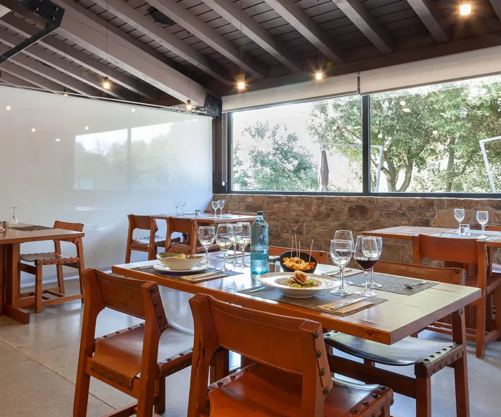 Comedor con mesa puesta, sillas de madera y ventanales con &aacute;rboles afuera