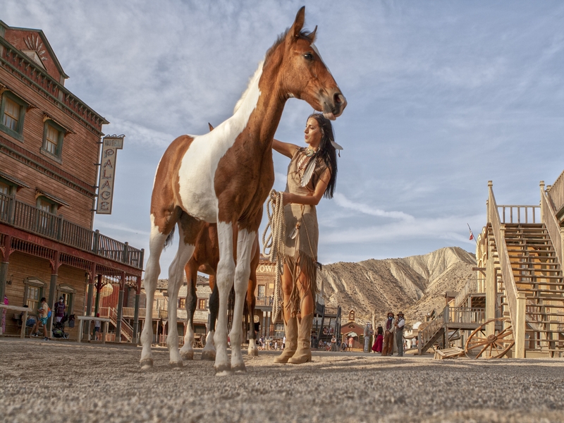 Plaza del Poblado de Oasys Minihollywood con una animadora disfrazada de india acariciando un caballo marrón y blanco. 