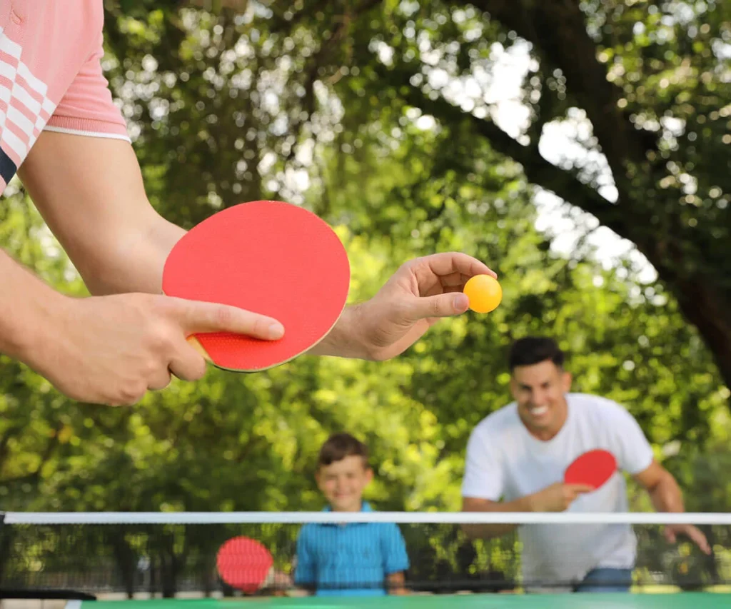Hombres jugando ping-pong con raquetas rojas y pelota amarilla en el parque