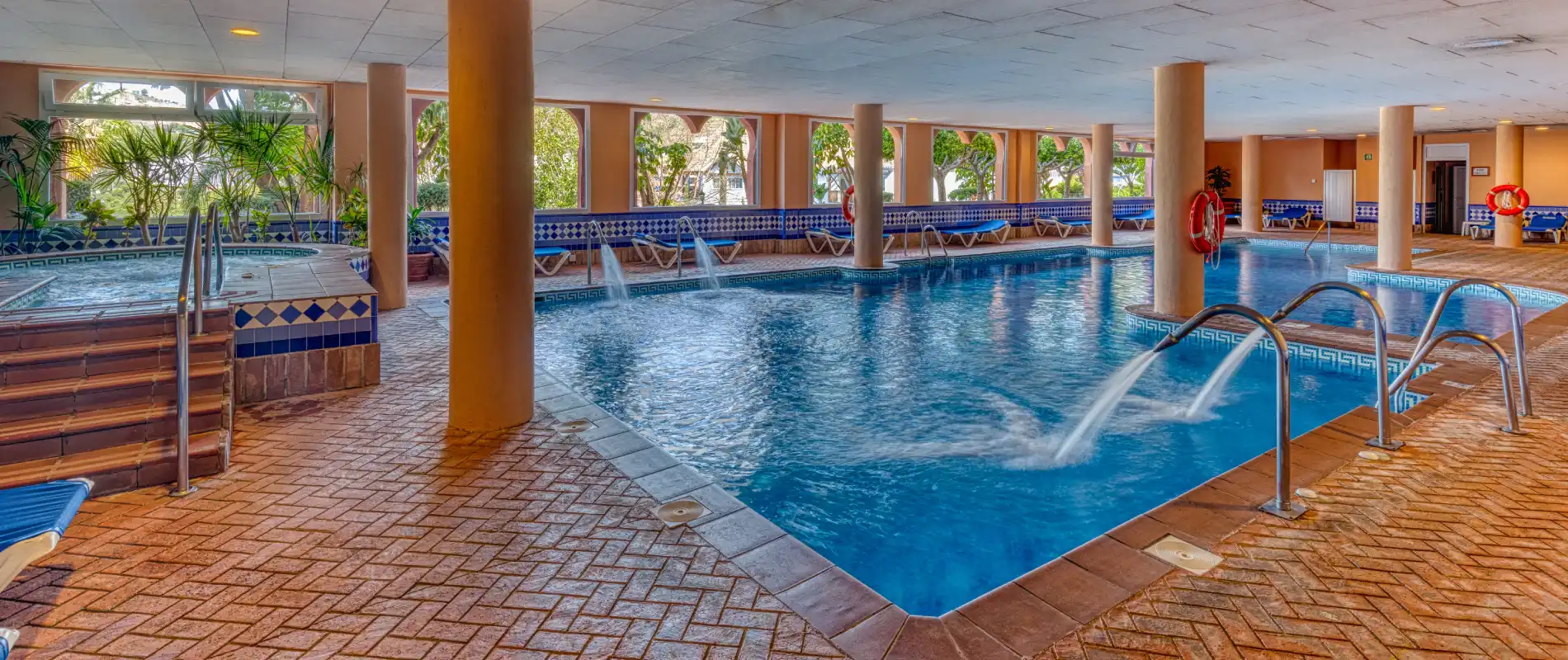 Piscina cubierta con chorros, columnas y tumbonas en un hotel.