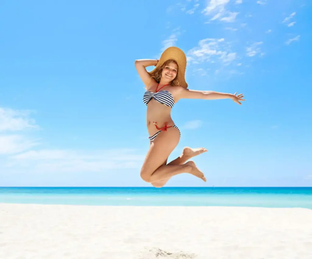 Ni&ntilde;a saltando en bikini con sombrero en la playa bajo cielo azul