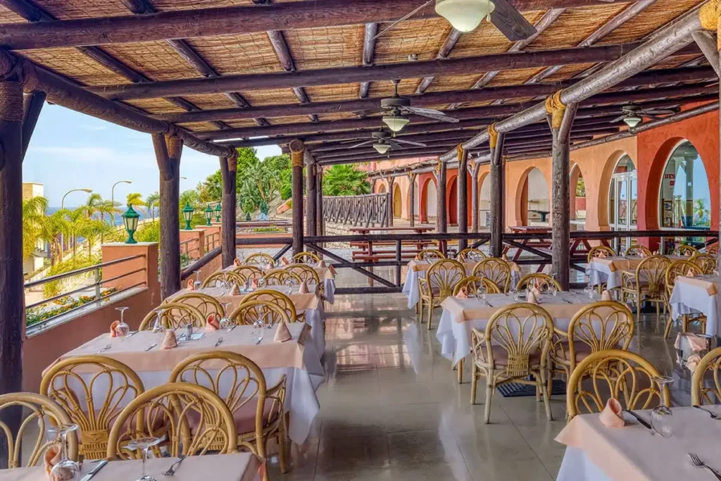 Terraza de restaurante con mesas, sillas de mimbre y techo de madera, junto al mar