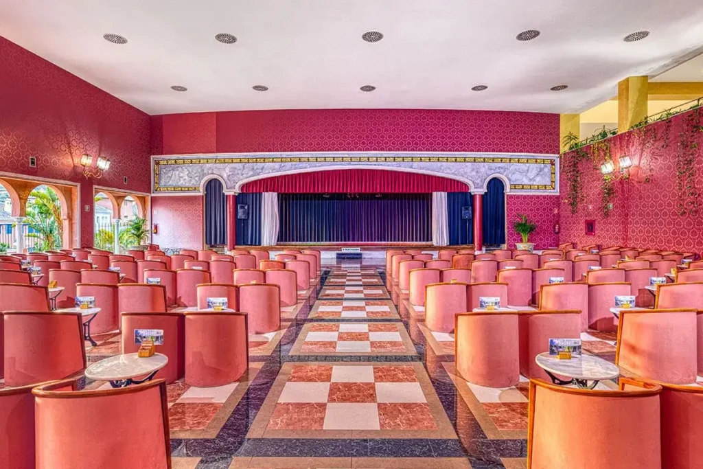 Sala de teatro con butacas rojas, pasillo central y escenario con cortinas.