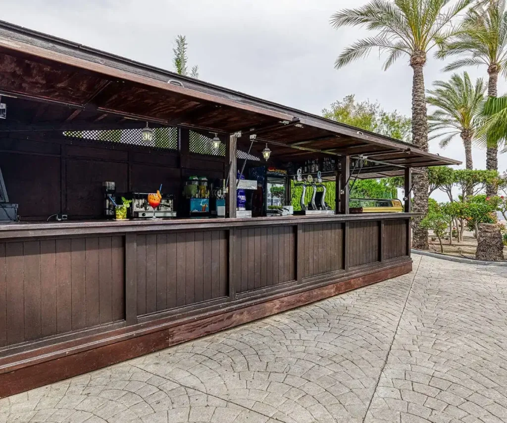Bar al aire libre con barra de madera, palmeras y mesas bajo techo.