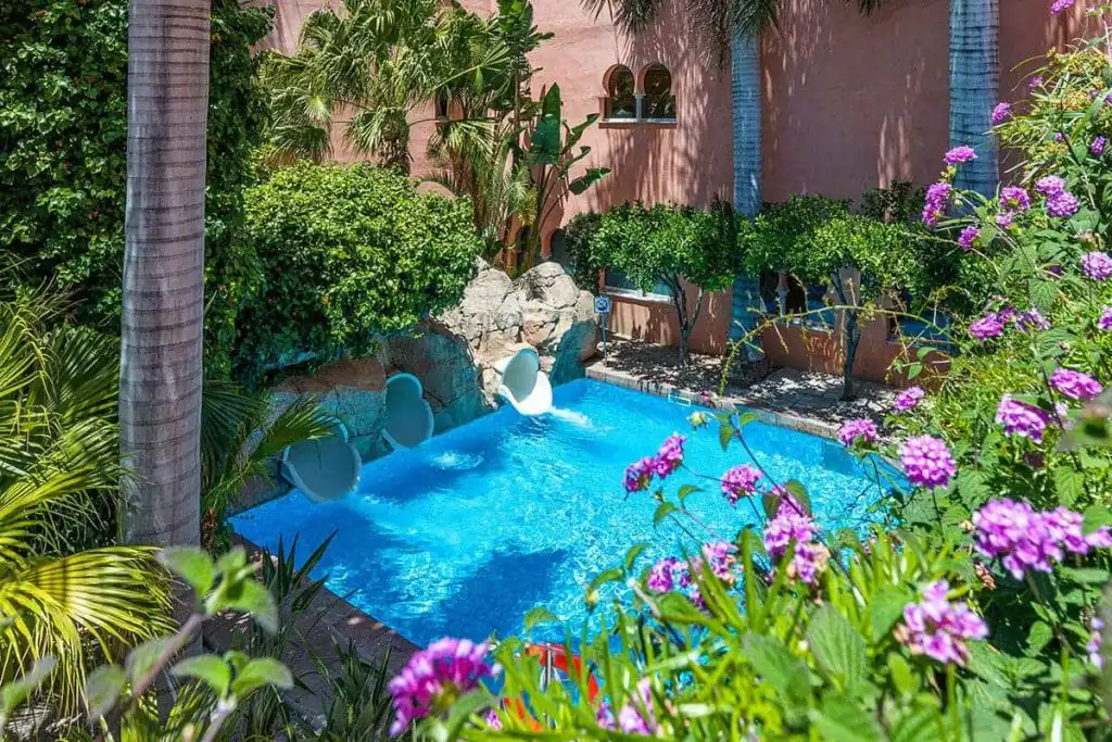 Piscina azul rodeada de palmeras y flores moradas en un jard&iacute;n tropical