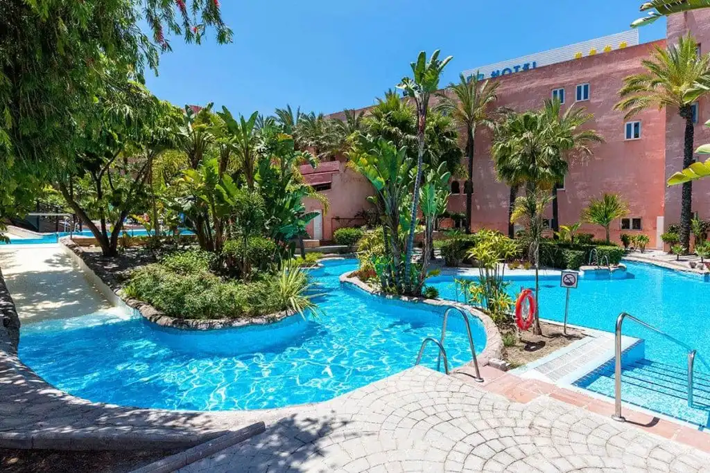 Piscina azul en resort tropical con palmeras y edificio rosado al fondo