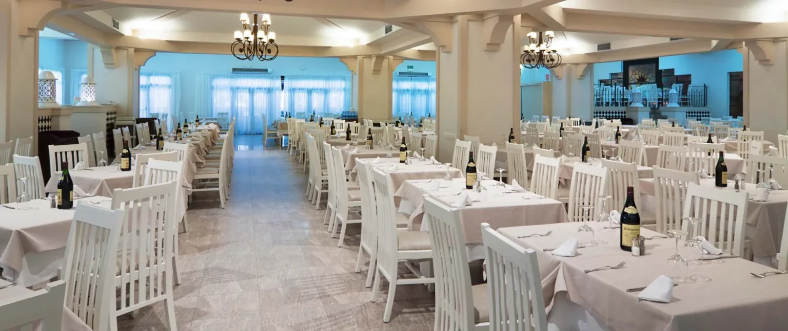Comedor de banquete con mesas rectangulares, sillas blancas y botellas de vino.