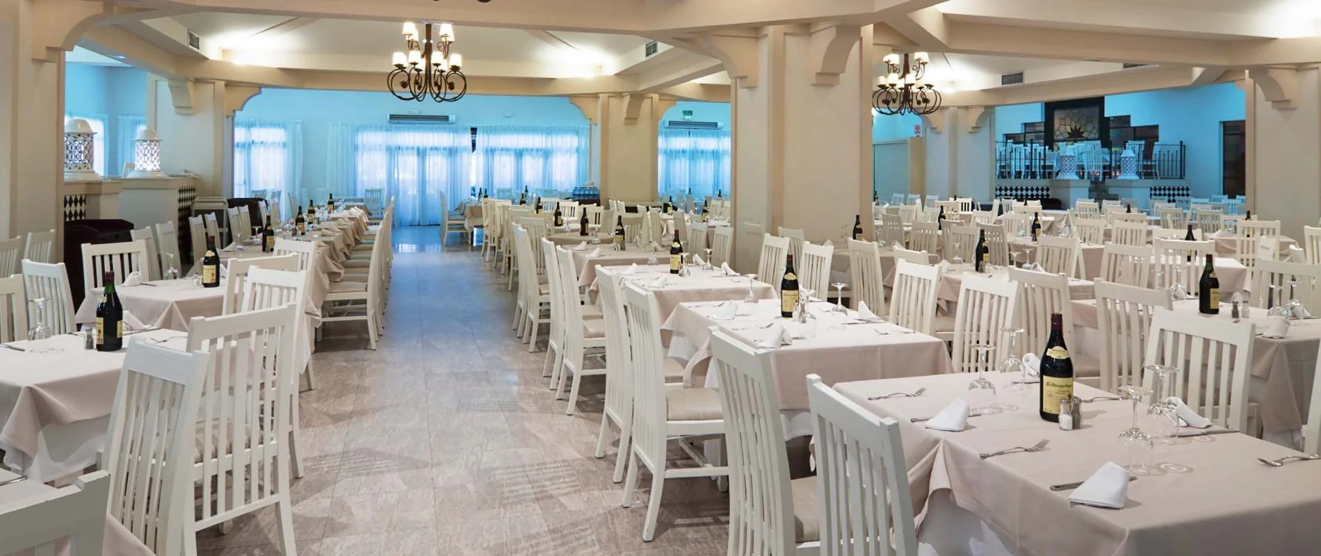 Comedor de banquete con mesas rectangulares, sillas blancas y botellas de vino.