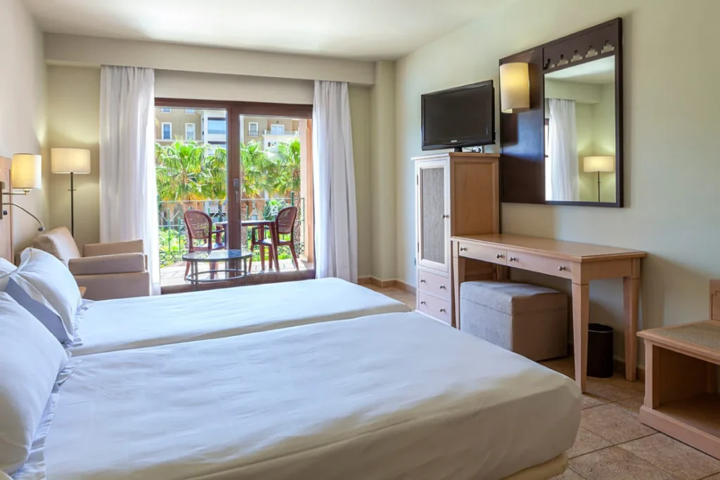 Habitaci&oacute;n de hotel luminosa con cama, escritorio y vista al balc&oacute;n y palmeras.