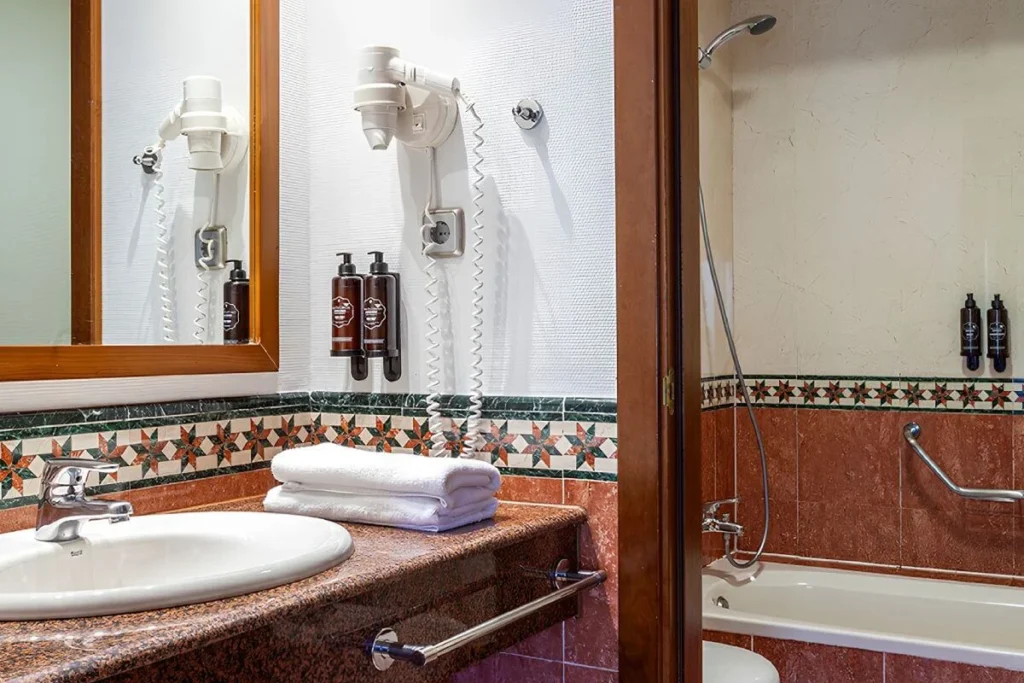 Ba&ntilde;o con lavamanos, toallas y dispensadores junto a ba&ntilde;era y espejo
