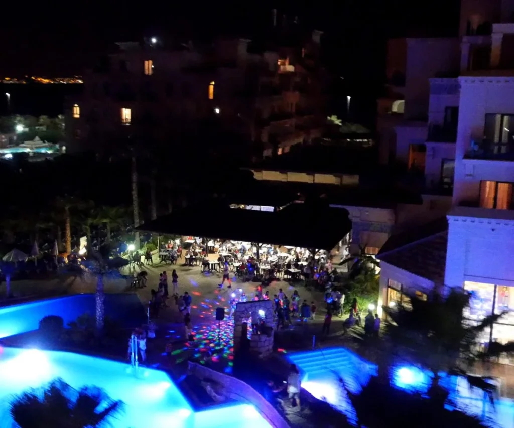 Vista nocturna de hotel con piscina iluminada, gente y evento al aire libre