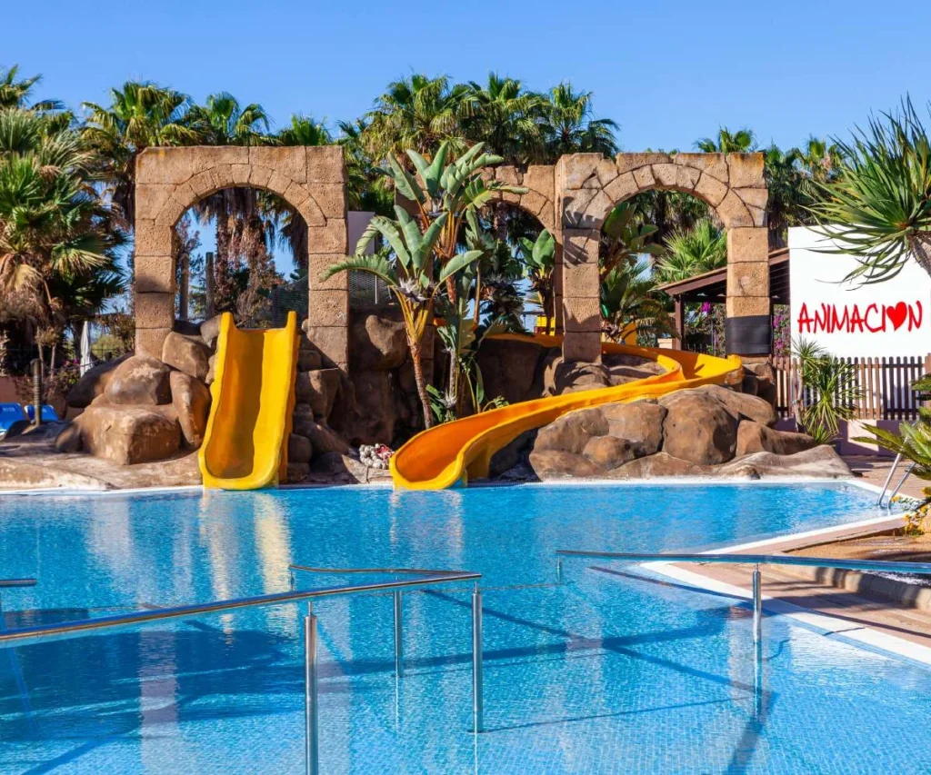 Piscina con toboganes amarillos y arcos de piedra entre palmeras, al sol