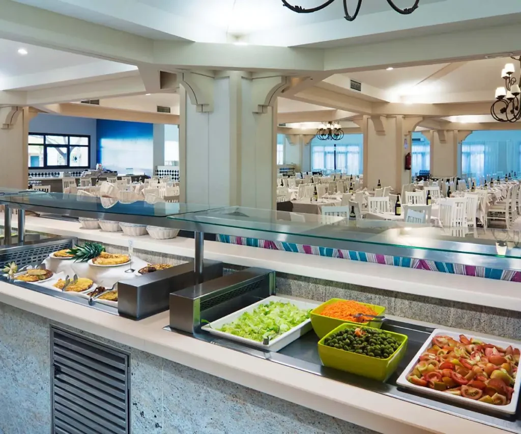 Buffet de alimentos con mesas, platos y bebidas en un sal&oacute;n interior con luz natural
