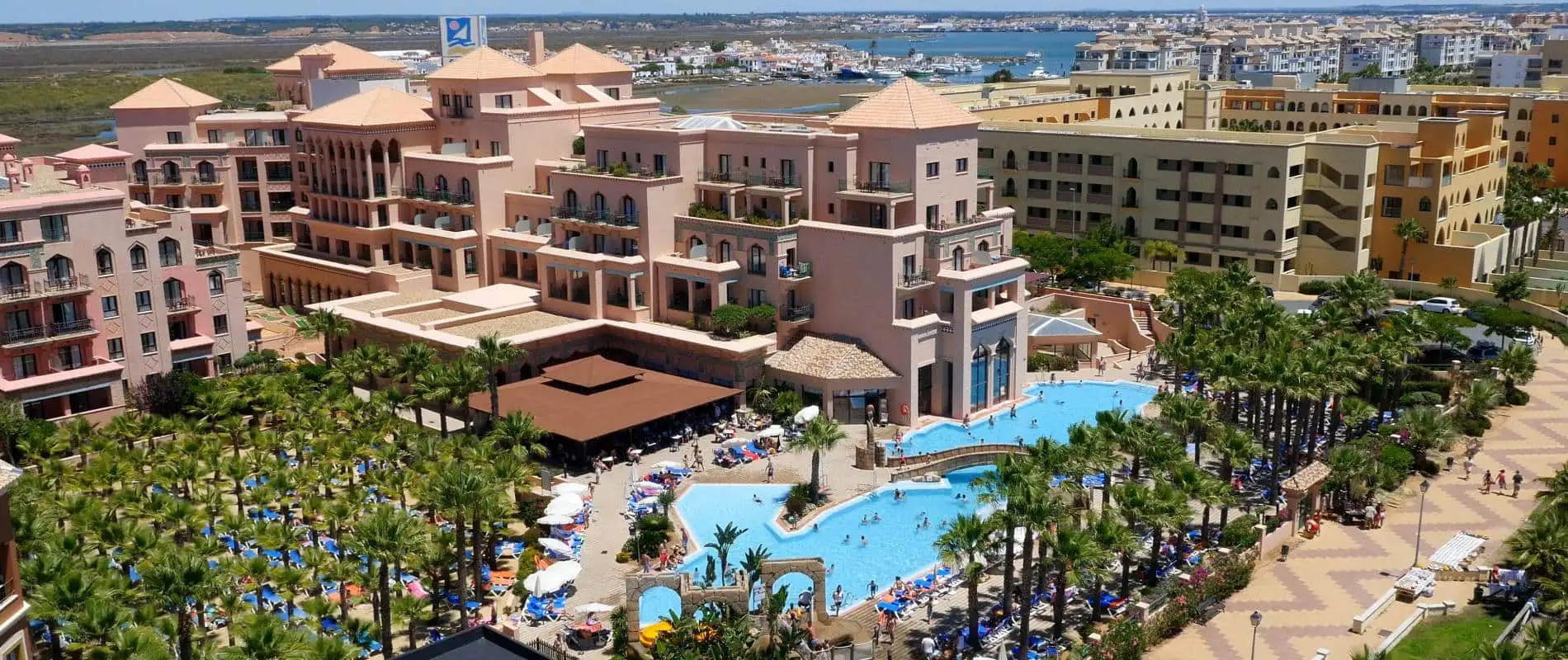 Vista a&eacute;rea de hotel con piscinas azules y palmeras junto al puerto y el r&iacute;o