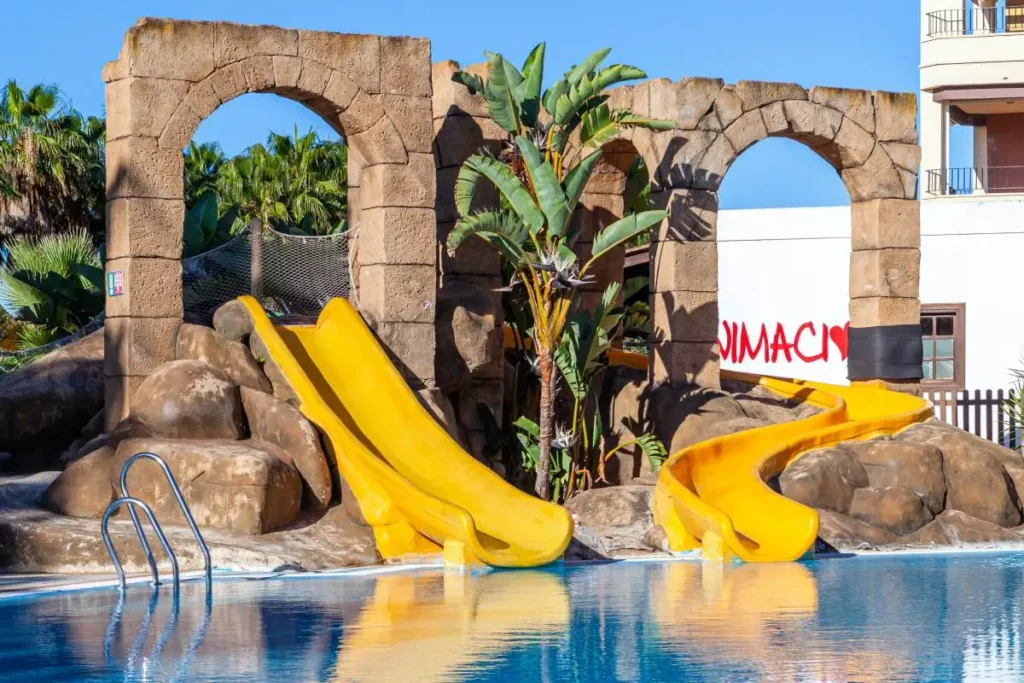 Arco de piedra y toboganes amarillos en piscina con palmeras y cielo azul