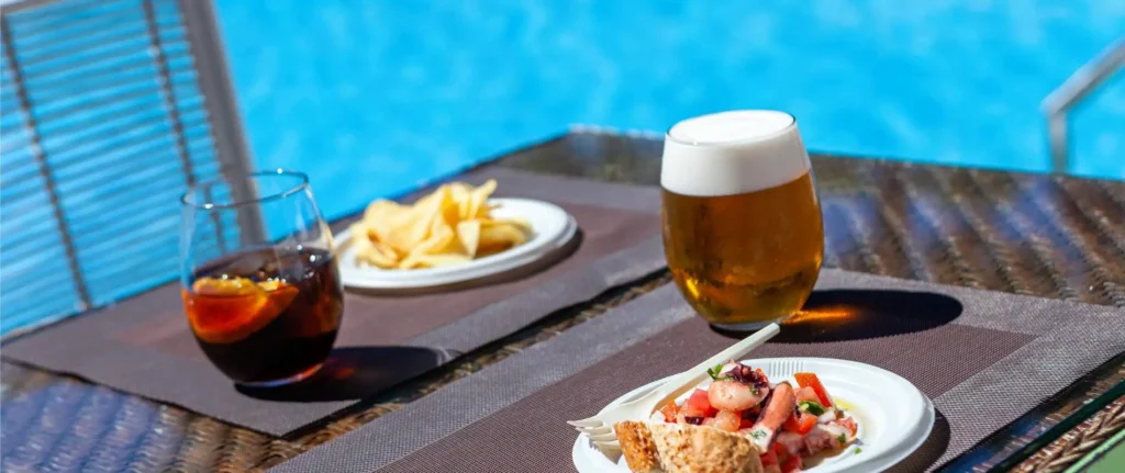Mesa con cerveza y refresco, acompa&ntilde;ados de comida en exterior junto a piscina.