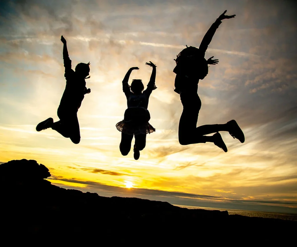 Tres siluetas saltando en un atardecer con cielo nublado y sol bajo