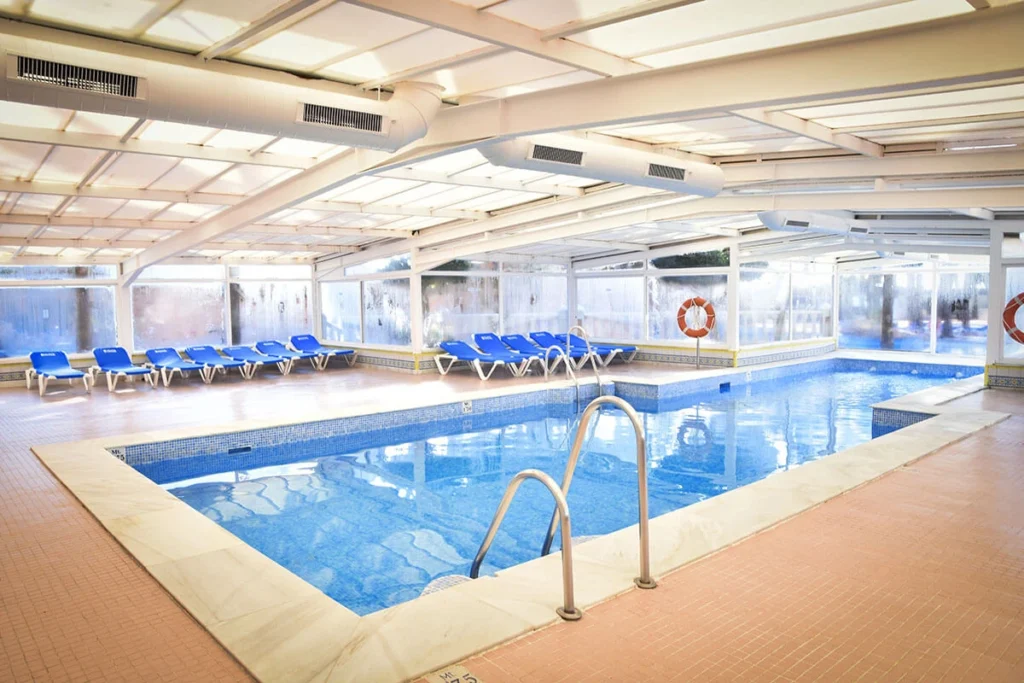 Piscina cubierta con tumbonas azules y ventanas al exterior, bajo techo blanco