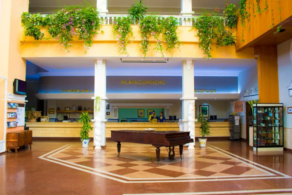 Vest&iacute;bulo hotelero con mostrador y piano, plantas colgantes y letreros.