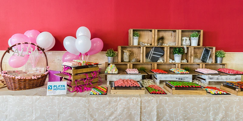Mesa decorada con dulces, globos rosas y elementos decorativos en pared roja.
