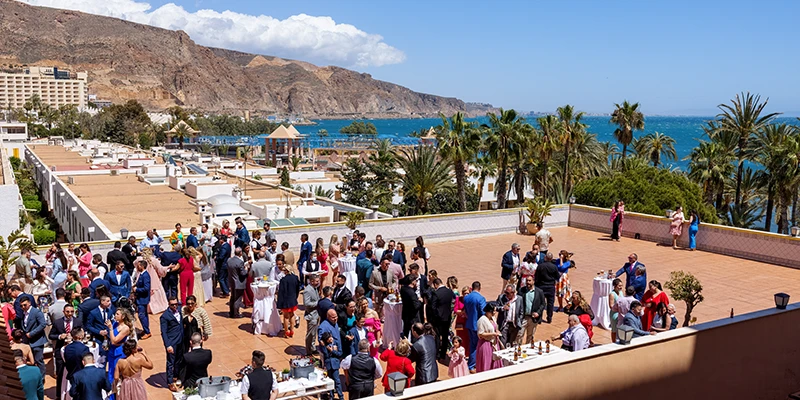 Grupo de personas en una terraza con vista al mar y monta&ntilde;as de fondo.